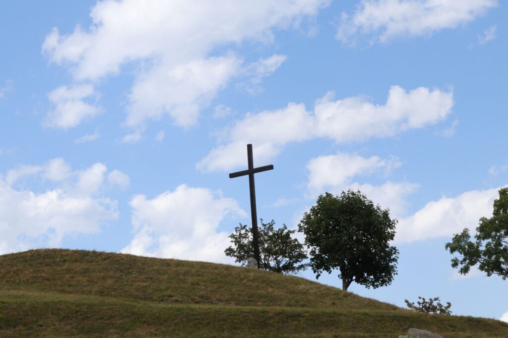 A silhouette of a cross on a grassy hill in Limonetto, Italy, with a bright blue sky and clouds